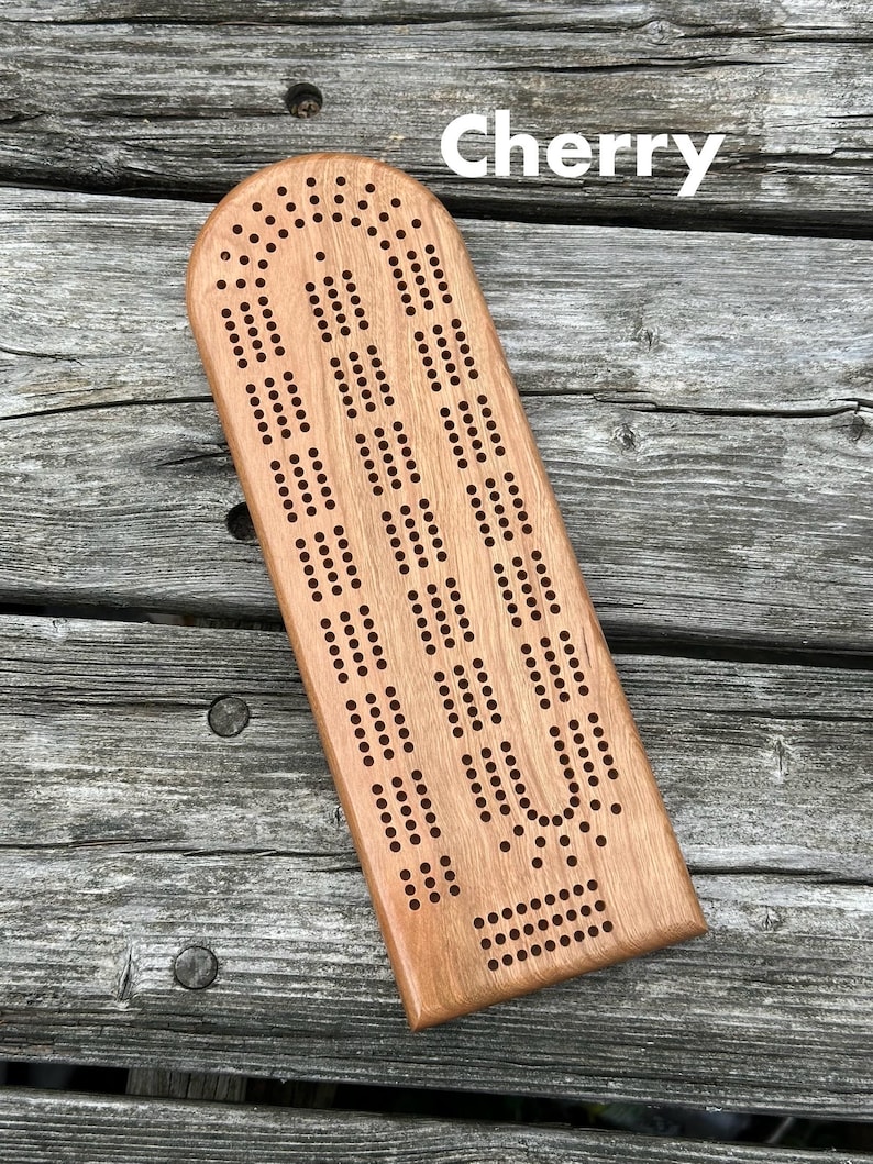 May include: A light brown cherry wood cribbage board with a rounded top edge and a continuous track of drilled holes for scoring. The word Cherry is printed in white text near the top of the board, which features a standard three-track layout.