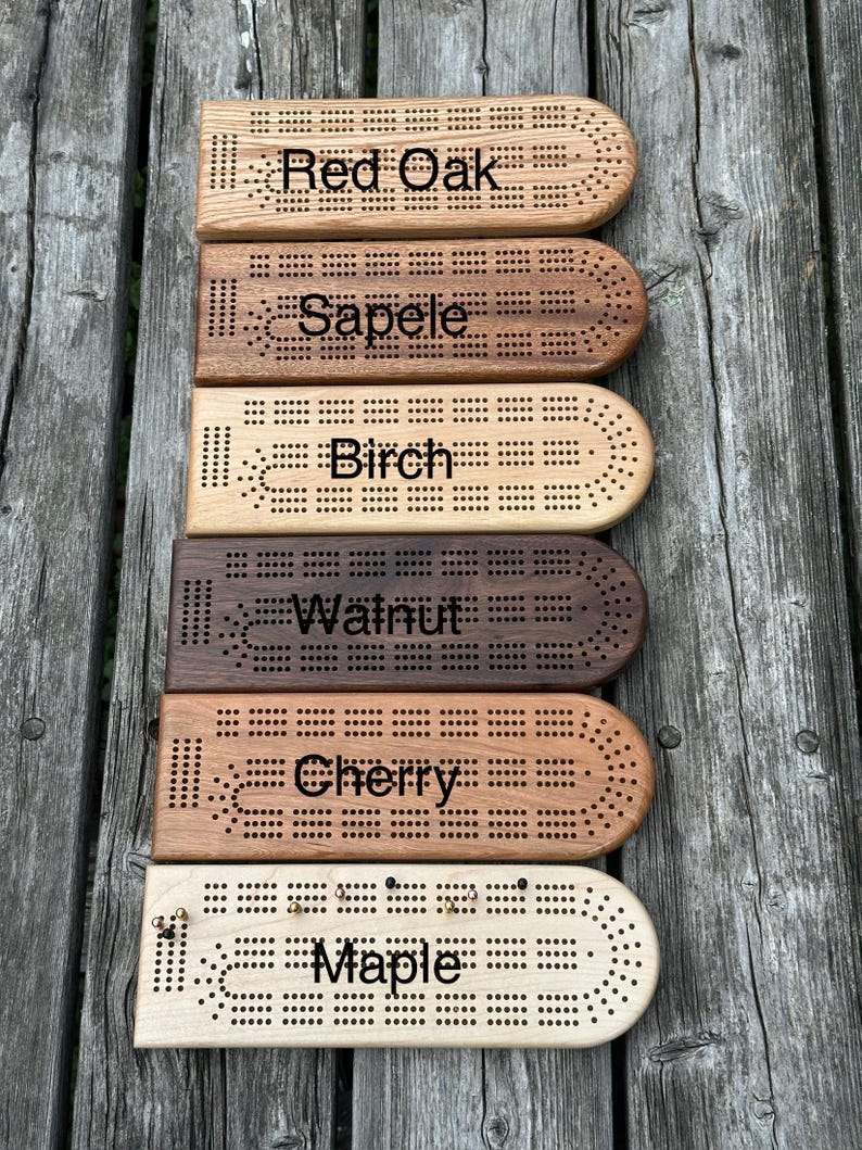 May include: Six rectangular wooden cribbage boards with rounded ends, each featuring drilled peg holes and black text labels identifying the wood types: Red Oak, Sapele, Birch, Walnut, Cherry, and Maple. The boards are arranged in a vertical stack.