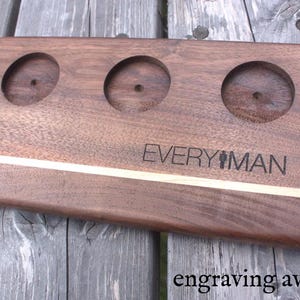 May include: A wooden serving tray with three circular cutouts and a light brown stripe across the top. The tray is engraved with the text "EVERYMAN" and two stick figures.