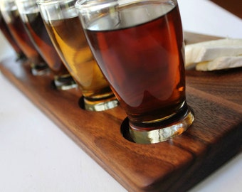 Puede incluir: Una bandeja de servir de madera con cuatro vasos llenos de bebidas de diferentes colores. La bandeja tiene un acabado de madera natural y está hecha de madera de nogal. También hay tres trozos de queso brie en la bandeja.
