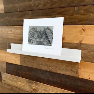 May include: A white shelf with a black and white photo of a church on it. The shelf is mounted on a wood wall.