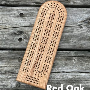 May include: A light brown red oak cribbage board with a rounded top edge and a rectangular base. The board features three parallel tracks of drilled holes for scoring, with a separate section of holes at the bottom for game tracking and peg storage.