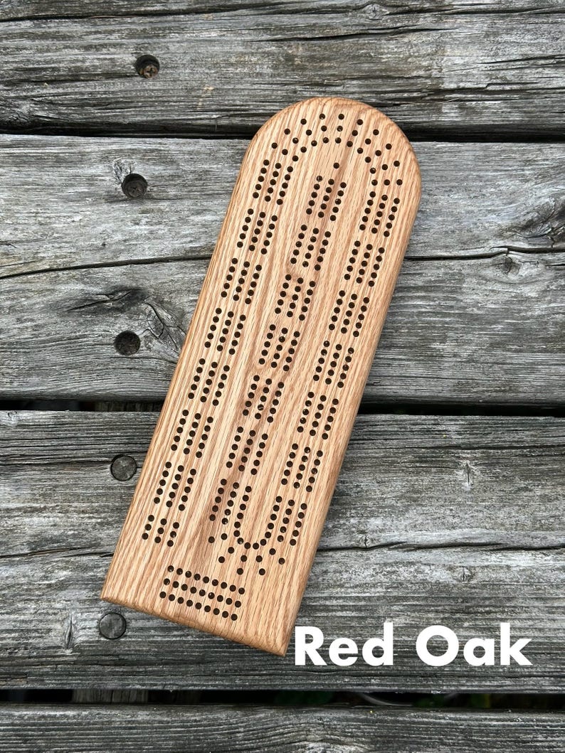 May include: A light brown red oak cribbage board with a rounded top edge and a rectangular base. The board features three parallel tracks of drilled holes for scoring, with a separate section of holes at the bottom for game tracking and peg storage.