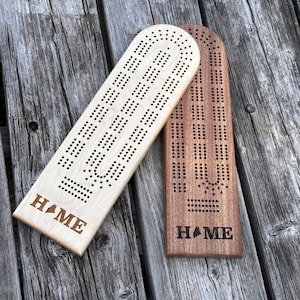Puede incluir: Dos tableros de cribbage de madera, uno claro y otro oscuro, se muestran sobre una superficie de madera envejecida. Cada tablero tiene una serie de agujeros perforados y la palabra "HOME" con un contorno estatal. Los tableros miden unos 30 cm de largo.