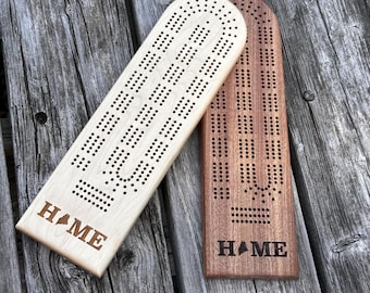May include: Two wooden cribbage boards with rounded tops, one light-colored and one dark-colored. Each board features a grid of drilled holes for scoring and the word HOME engraved at the bottom, with the letter O replaced by a map of Maine.