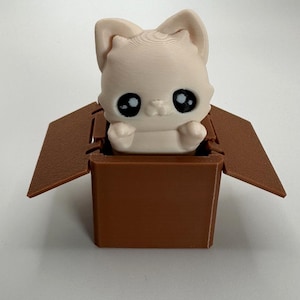 Puede incluir: Una pequeña figura de gato blanca impresa en 3D con grandes ojos negros se sienta dentro de una caja de cartón marrón con la tapa abierta.