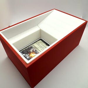 May include: A red and white card storage box with a white interior and a single card displayed in the opening. The card is a Magic: The Gathering card with a white border and a colorful illustration.