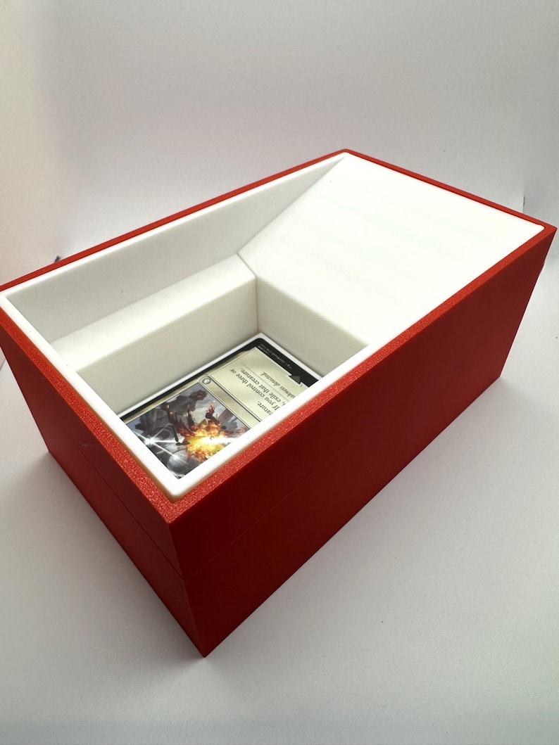 May include: A red and white card storage box with a white interior and a single card displayed in the opening. The card is a Magic: The Gathering card with a white border and a colorful illustration.