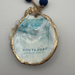 May include: An oyster shell ornament with a gold-painted edge, featuring a painted coastal scene in shades of blue and white. The word "SOUTHPORT" is printed on the shell. It is suspended by a twine loop with two blue beads.