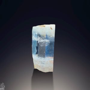 Puede incluir: Cristal de aguamarina rectangular y translúcido, de color azul claro. El cristal presenta una textura natural y rugosa, con una superficie irregular. La base del cristal es de un blanco roto pálido, que se transforma en un azul claro transparente hacia la parte superior.
