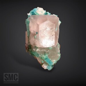 May include: A natural mineral specimen featuring a large, light pink crystal with a geometric shape. The crystal is accented with teal and green mineral formations, creating a striking contrast. The overall composition is set against a dark background.