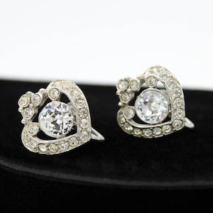 以下が含まれることがあります： ハート型のシルバーイヤリングのペア。各ハートの中央に大きなクリアラインストーンがセットされています。イヤリングは小さなラインストーンで囲まれています。