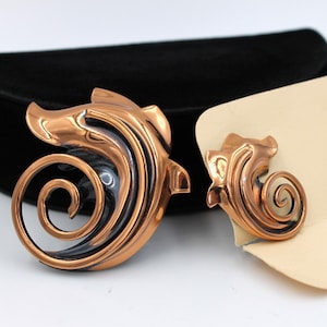 May include: A set of copper earrings and a brooch with a swirling, abstract design. The jewelry is on a white card with the word "Ramé" printed on it.
