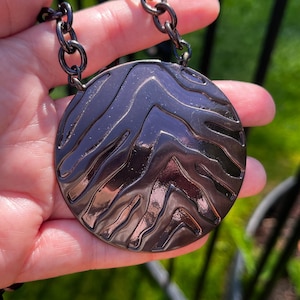 May include: A dark metallic gunmetal necklace featuring a large circular pendant with a raised, wavy, zebra-like striped texture. The pendant is suspended from a thick, matching dark metal chain link necklace with a polished, reflective finish.