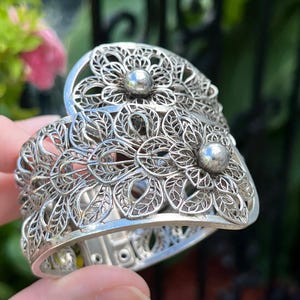 May include: Silver filigree cuff bracelet with an open floral design. The bracelet has two large flower-shaped accents, each with a small silver ball in the centre. The bracelet is held in a hand.