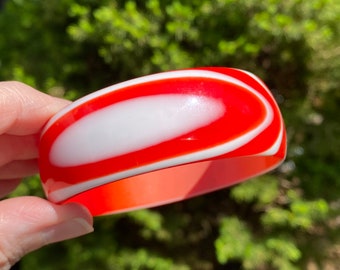 May include: A red and white plastic bangle bracelet with a swirl pattern. The bracelet is glossy and has a smooth finish.