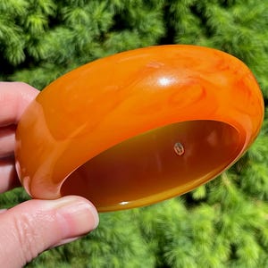 Könnte beinhalten: Ein großes, orange-bernsteinfarbenes Armreif-Armband. Das Armband hat eine glatte, glänzende Oberfläche mit marmoriertem Aussehen. Das Armband wird vor einem Hintergrund aus grünem Laub gehalten.
