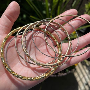 Puede incluir: Un conjunto de brazaletes de oro y plata, apilados y dispuestos en una mano. Los brazaletes son finos y delicados, con una variedad de texturas y acabados.