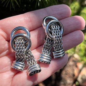 May include: A pair of silver tone earrings with a chain link design and a circular detail. The earrings are dangling and have a knotted design.