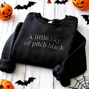 Puede incluir: Sudadera negra con la frase "A little ray of pitch black" bordada en la parte delantera. La imagen está decorada con elementos temáticos de Halloween, como calabazas y siluetas de murciélagos.