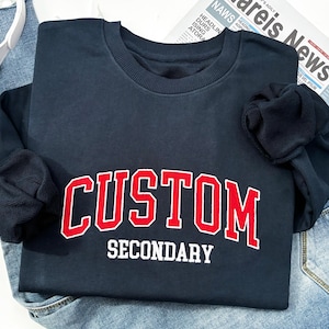 May include: A navy blue sweatshirt with the word "CUSTOM" in red block letters outlined in white, and "SECONDARY" in white block letters. The sweatshirt is folded on top of blue jeans. A pair of white headphones and a baseball cap are visible.
