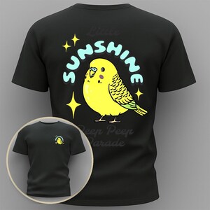 Puede incluir: Camiseta negra con una ilustración de periquito amarillo y el texto "Little Sunshine Peep Peep Parade". El diseño incluye texto azul y detalles de estrellas. Una versión más pequeña del diseño está en la parte delantera de la camiseta.