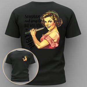 以下が含まれることがあります： 黒いTシャツ。ナイフを持った女性のレトロなイラストと「Sensitive and fragile but will still fight」というテキストが特徴です。デザインの小さなバージョンが前面にあります。