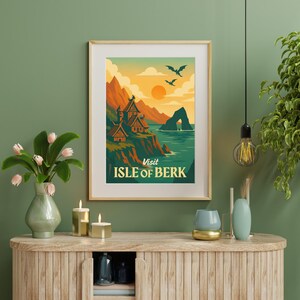 May include: A framed art print featuring a scenic landscape with the text "Visit Isle of Berk." The artwork depicts a coastal scene with mountains, buildings, and flying creatures, set against a sunset sky. The print is in a wooden frame.