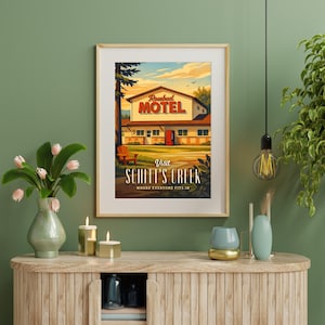 May include: Framed art print featuring a vintage-style illustration of the Rosebud Motel from Schitt's Creek. The artwork depicts the motel with a red door, a wooden chair, and the text "Visit Schitt's Creek". The print is in a wooden frame.
