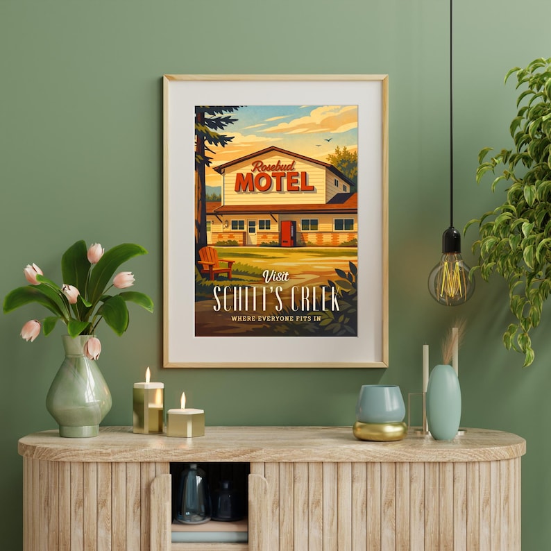 May include: Framed art print featuring a vintage-style illustration of the Rosebud Motel from Schitt's Creek. The artwork depicts the motel with a red door, a wooden chair, and the text "Visit Schitt's Creek". The print is in a wooden frame.