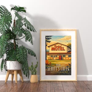 May include: A framed poster with a vintage travel-style illustration of the Rosebud Motel from Schitt's Creek. The poster features the motel building, a wooden chair, and the text "Visit Schitt's Creek Where Everyone Fits In."