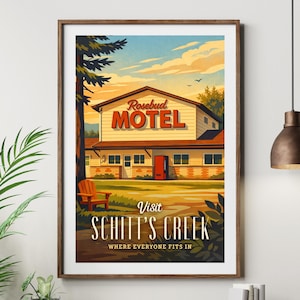 May include: A framed art print featuring the Rosebud Motel from Schitt's Creek. The motel is beige with a red door and the word "MOTEL" in red. The print includes the text "Visit Schitt's Creek" and a wooden chair.