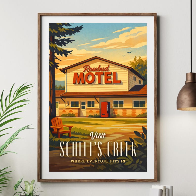 May include: A framed art print featuring the Rosebud Motel from Schitt's Creek. The motel is beige with a red door and the word "MOTEL" in red. The print includes the text "Visit Schitt's Creek" and a wooden chair.