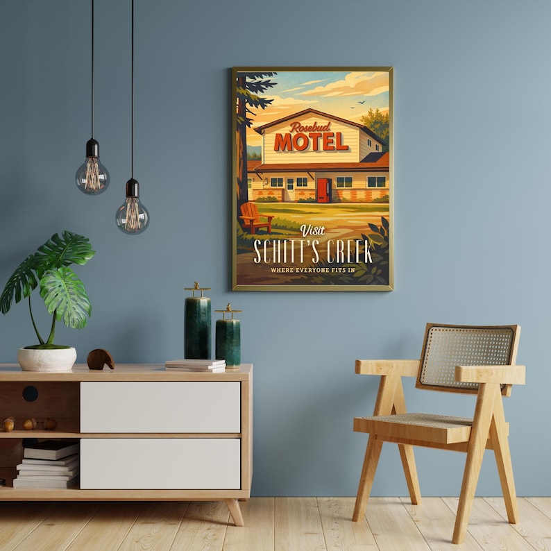 May include: A framed poster featuring the Rosebud Motel from Schitt's Creek. The artwork depicts the motel with a red sign, a sunny sky, and the text "Visit Schitt's Creek, Where Everyone Fits In."