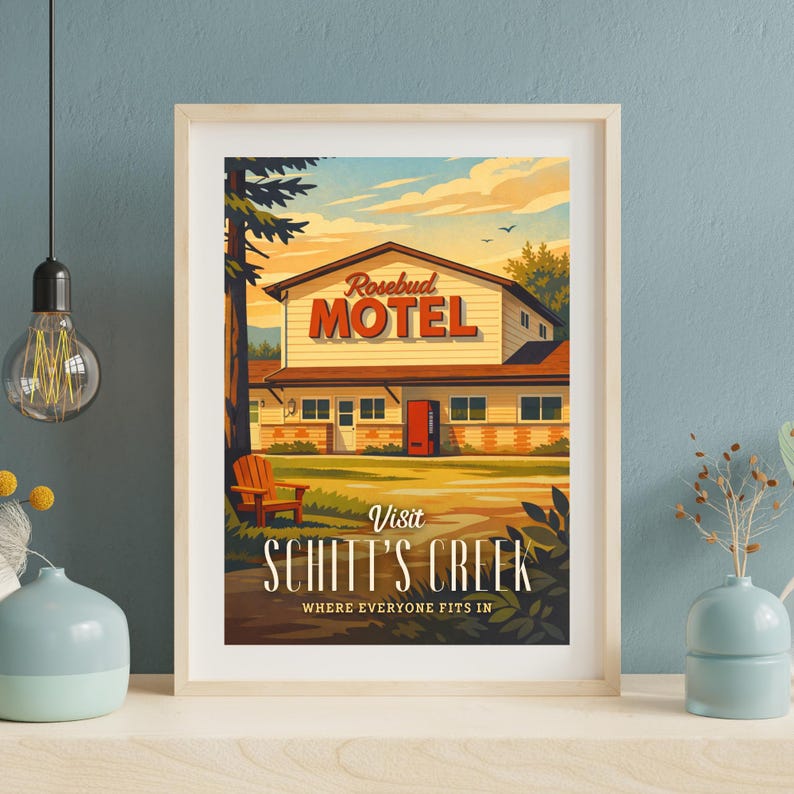 May include: Framed art print featuring the Rosebud Motel from Schitt's Creek. The print shows the motel with a red door, a wooden chair, and the text "Visit Schitt's Creek, Where Everyone Fits In."