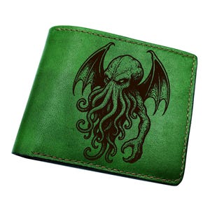 May include: Green leather wallet featuring a detailed brown illustration of a creature with wings and tentacles. The wallet has a bi-fold design with visible stitching around the edges. The overall aesthetic is a blend of fantasy and gothic themes.