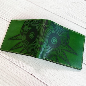 May include: Green leather wallet featuring a black Majora's Mask design. The wallet is open, revealing the interior. The edges are stitched with green thread. The wallet is on a light wood surface.