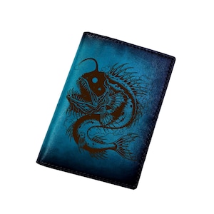 Peut inclure: Un porte-passeport rectangulaire en cuir avec un dégradé bleu profond, orné d'une illustration sombre et détaillée gravée au laser représentant une baudroie avec des dents acérées et un leurre lumineux, doté de bords cousus et d'un design fin.
