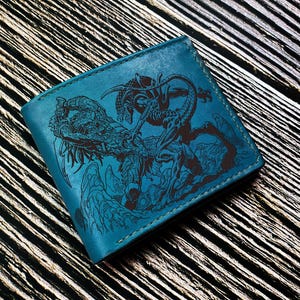 May include: A teal leather wallet featuring a detailed black illustration of a creature. The wallet is rectangular and appears to be made of high-quality leather. The design is intricate and the color contrast is striking.