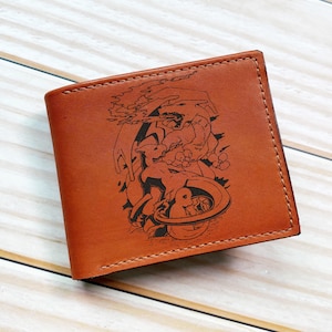 May include: A brown leather wallet with a black graphic design. The wallet is rectangular and has a stitched border. The graphic appears to be a cartoon illustration of a character. The wallet is closed and sits on a light wood surface.