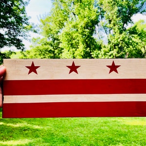 Op de afbeelding: Een houten bord geschilderd met de vlag van Washington, D.C. De vlag heeft drie rode sterren op een witte achtergrond met twee rode horizontale strepen.