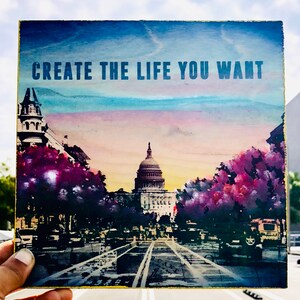 May include: A wooden sign with a blue, pink, and yellow watercolor background. The sign says "CREATE THE LIFE YOU WANT" in bold, blue letters. The sign features a cityscape with a large building in the center.