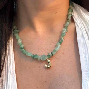 Peut inclure: Collier de pierres vertes irrégulières avec un pendentif doré en forme de lettre "J". Le collier est composé de petites pierres vertes. Le pendentif est une lettre "J" stylisée. Le collier est porté autour du cou.