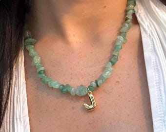 May include: A green stone necklace with a gold-colored initial "J" pendant. The necklace is made of small, irregularly shaped green stones. The pendant is a stylized letter "J". The necklace is worn around the neck.