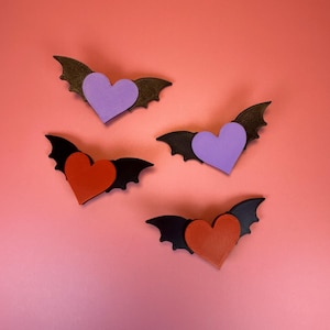 May include: Four bat-shaped decorations with heart-shaped centers. Two have purple hearts and brown wings, while the other two have red hearts and black wings. The decorations are arranged on a pink background.