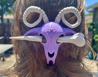 May include: A purple and white hair accessory with a ram skull design. The accessory has curved white horns, a bone-shaped pin, and a detailed skull. The hair piece is worn in the hair.