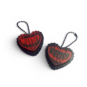 May include: Two heart-shaped keychains with a black scalloped edge and a red center. Each keychain features the word MURDER in raised black lettering, surrounded by a black spiderweb design, and includes a black ball chain attachment.