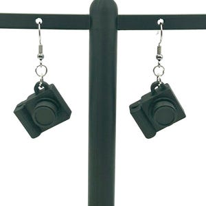 May include: A pair of black camera-shaped earrings hanging from a dark green display stand. The earrings have a detailed design, including a lens and buttons. The earrings are suspended by silver-colored hooks.