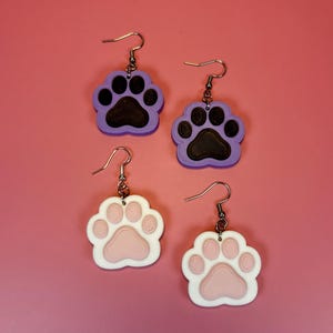 May include: Four pairs of paw print earrings. Two pairs have purple and black paw prints, and two pairs have white and pink paw prints. The earrings are hanging on silver hooks against a pink background.