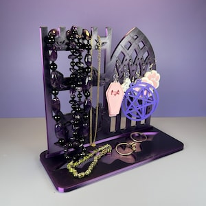 May include: Purple jewelry display stand with a gothic arch design. The stand holds necklaces, earrings, and rings. Jewelry includes black beaded necklaces, a gold chain, and earrings with coffin and pentagram charms. The stand is on a purple base.